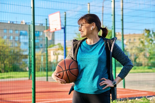 Middle aged smiling active woman with ball, near the outdoor basketball playground court. Active leisure, urban sport, healthy lifestyle, mature people concept