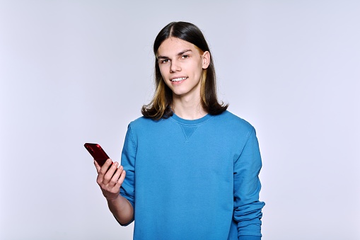 Handsome guy with smartphone in his hand, on light studio background. Young positive male student 17, 18 years old looking at camera. Youth, technology, lifestyle, communication, young people concept