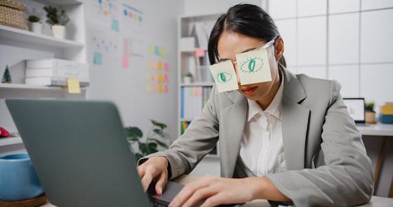 Young Asian businesswoman sitting on desk overworked tired sleep over a laptop at office. Exhausted burnout lady  with two post-it over her eyes, adhesive notes on face sleeping at workplace.