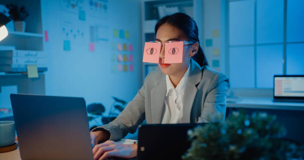 Young Asian businesswoman sitting on desk overworked tired sleep over a laptop at office at night. Young Asian businesswoman sitting on desk overworked tired sleep over a laptop at office at night. Exhausted burnout lady with two post-it over her eyes, adhesive notes on face sleeping at workplace. wasting time stock pictures, royalty-free photos & images