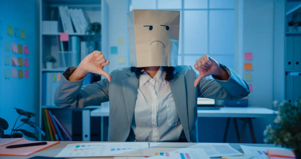 Young Asian businesswoman sit on desk wear draw sad mask paper craft bag feel bad mood with fail work project in office at night. stock photo