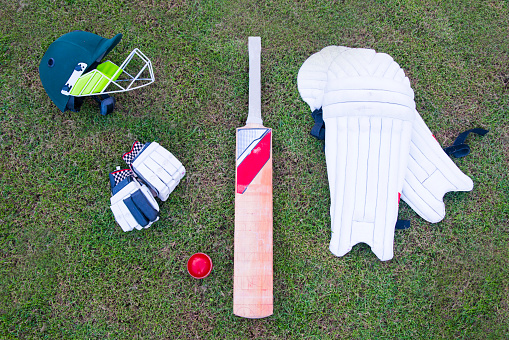 Flat lay image of cricket equipment laying on the grass. Consists of batters helmet, cricket ball, cricket bat, and batters pads.