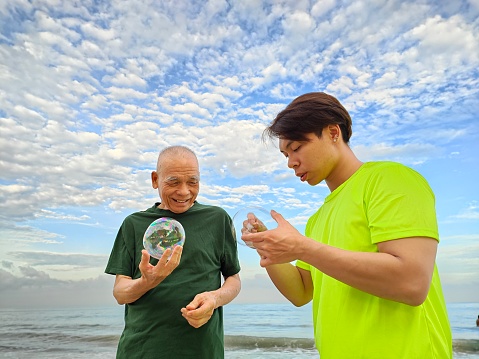 An Asian senior man is learning to blow bubble with his grandson by the beach.