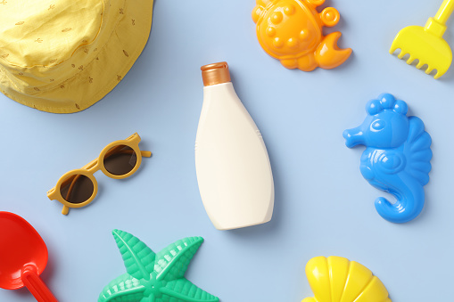 Baby sunscreen lotion with sunglasses, sand molds and panama hat on blue table. Flat lay, top view.