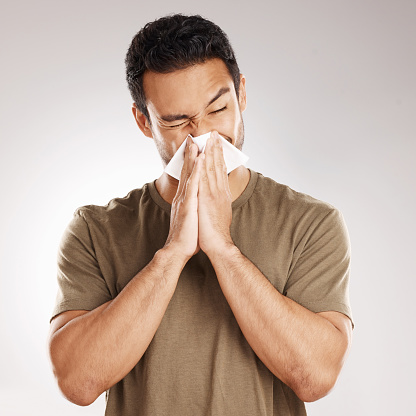 Handsome young mixed race man blowing his nose while standing in studio isolated against a grey background. Hispanic male suffering from cold, flu, sinus, hayfever or corona and using a facial tissue