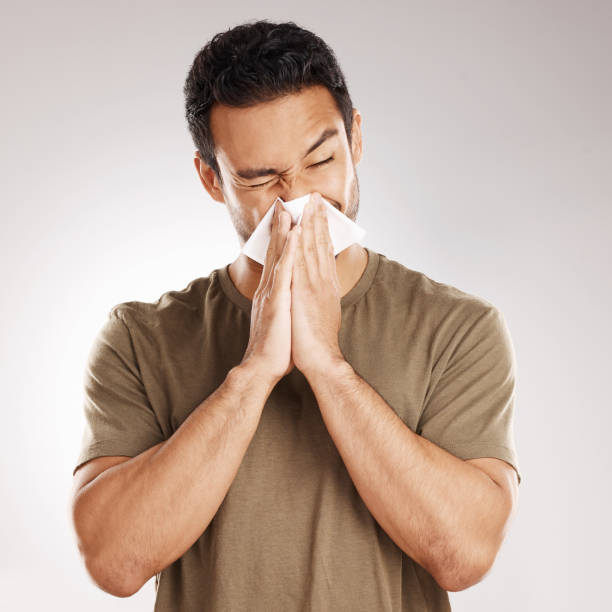 handsome young mixed race man blowing his nose while standing in studio isolated against a grey background. hispanic male suffering from cold, flu, sinus, hayfever or corona and using a facial tissue - resfriado e gripe imagens e fotografias de stock