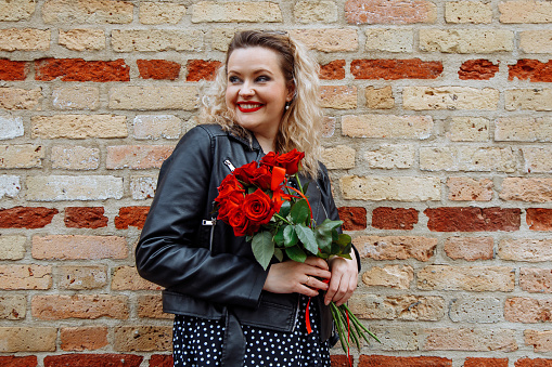 Stylish, confident embarrassed blond woman with make-up, posing in leather jacket and dress against brick colorful wall with bouquet roses in hands. City street style. Birthday event, congratulation