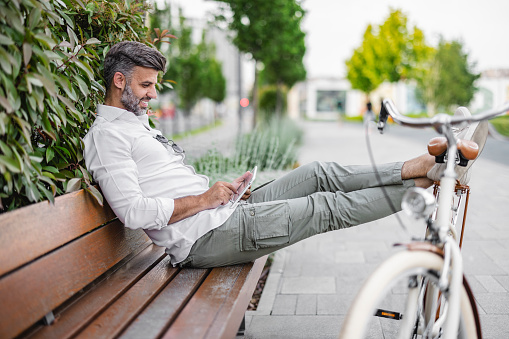 Handsome commuter working on tablet computer while sitting on a park bench next to a bicycle