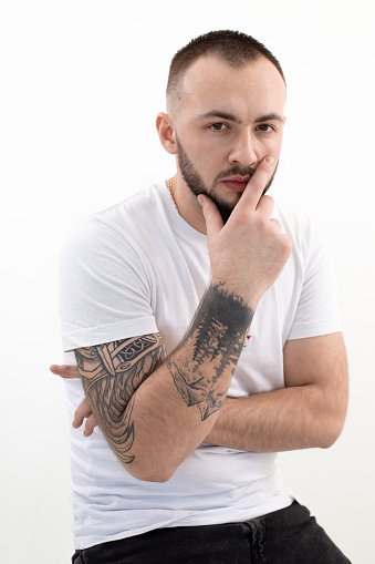 Vertical shot of tattooed brunet, bearded confident man in casual clothes thinking in studio on white background. Smart, confident businessman, brainstorming at working process. Holding chin by hand