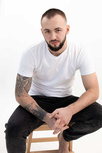 Vertical photo of tattooed brunet, bearded confident man in casual clothes thinking in studio on white background. Smart, confident businessman, brainstorming at working process. Cross arms on legs