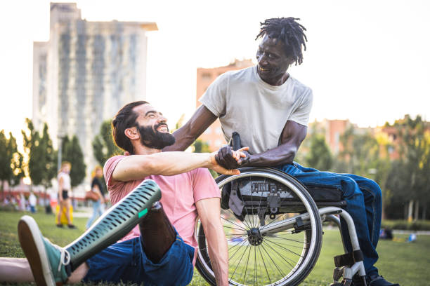 deux amis qui ont des handicaps physiques différents se saluent au parc, un homme africain adulte en fauteuil roulant serrant la main de son ami hispanique avec une jambe artificielle - prosthetic equipment photos et images de collection