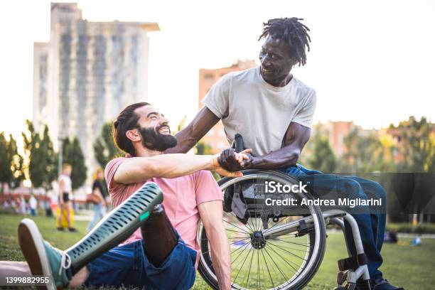 Two Friends Who Have Different Physical Disabilities Greet Each Other At The Park Adult African Man In Wheelchair Shaking Hands With His Hispanic Friend With An Artificial Leg Stock Photo - Download Image Now