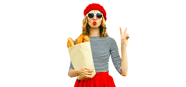 Portrait of beautiful woman blowing her red lips sending sweet air kiss holding grocery shopping paper bag with long white bread baguette wearing french red beret isolated on white background