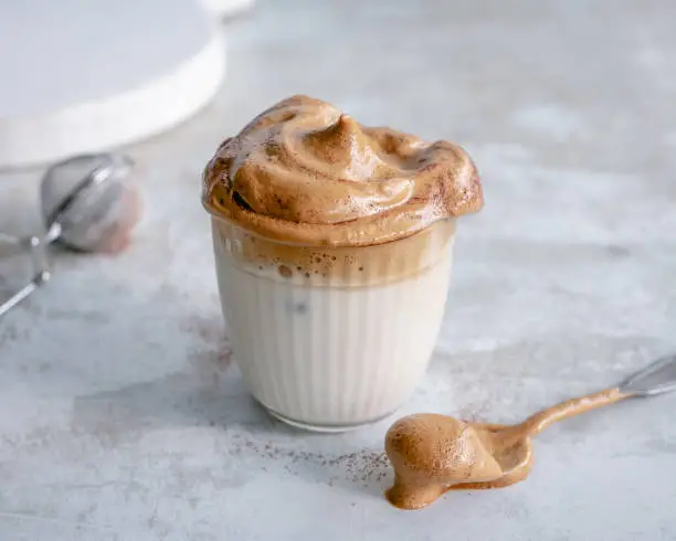 Glass of dalgona coffee sprinkled with cocoa powder and a spoon next to it, shot on a minimal background