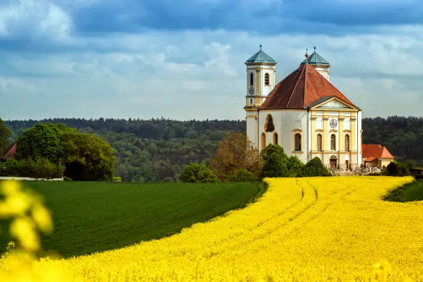 Germany, Bavaria, Burghausen. Marienberg Church surrounded by a rapeseed field. The Marienberg pilgrimage church is called the "Pearl of the Salzach Valley". When the Cistercians from Schützing moved their monastery to Raitenhaslach, there was a "Capella" in Marienberg. Over the centuries, the church has been rebuilt, expanded or - most recently in 1760 - rebuilt.