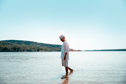 Man walking at the beach on sunny day. Handsome man in white shirt and sun hat walk through shallow water. Copy space