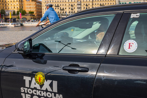 Sweden. Stockholm. 05.18.2022. Close up view of driver in Taxi Stockholm vehicle and cyclist on old buildings background.