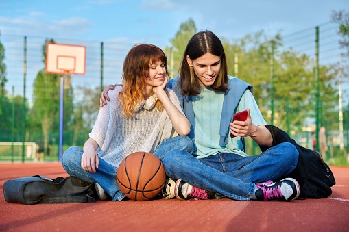 Couple of teenage student friends sitting on outdoor basketball court, with ball backpacks, laughing, talking, looking in smartphone. Youth, active healthy lifestyle, education, urban sport concept