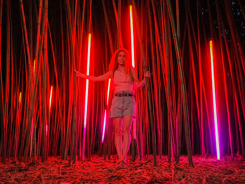 A woman stands in night bamboo grove among the trunks that sparkle in red. Walk through the southern resort