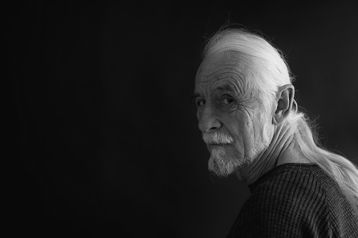 Low key studio black and white portrait of beautiful old man with gray hair in a braid looking over his shoulder at the camera. Horizontally.