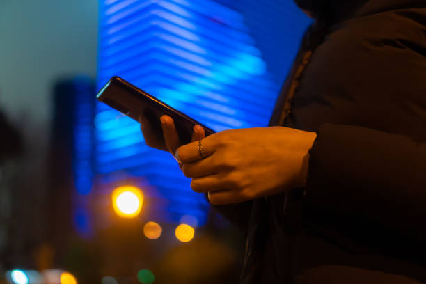 Close-up of a woman's hands with a mobile phone at night on the street against the background of a lighted blue building, copy space Close-up of a woman's hands with a mobile phone at night on the street against the background of a lighted blue building, copy space Iphone stock pictures, royalty-free photos & images