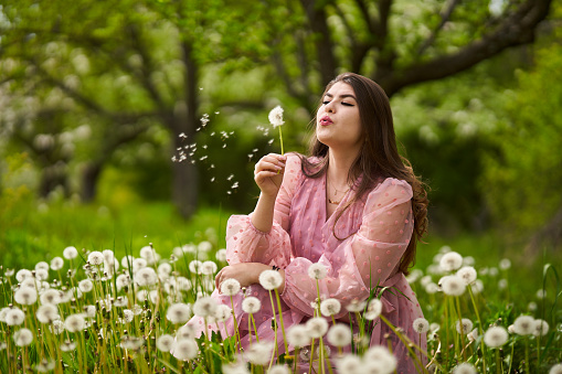 Young woman in an orchard, blowing dandelion seeds in the wind