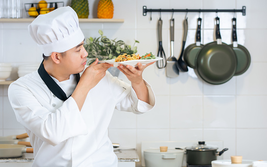 Portrait beautiful Asian professional male chef wearing white uniform, hat, holding, showing plate of spaghetti, cooking in kitchen, making surprising face with happiness. Restaurant, Food Concept