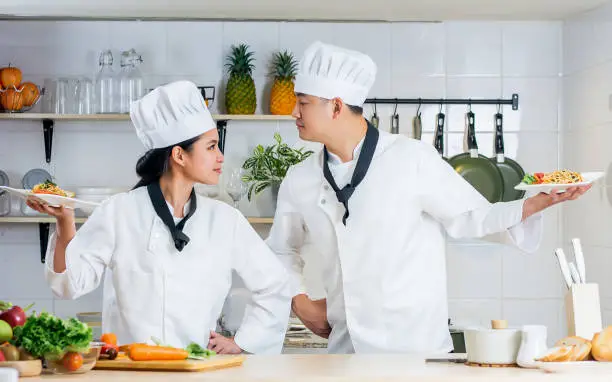 Two Asian professional chef wearing white uniform, hat, holding plates of spaghetti, cooking, fighting in kitchen, looking each with confidence other for competition in contest. Food Concept