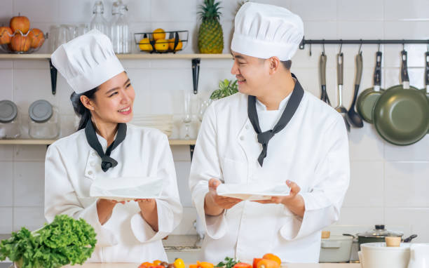istock Two Asian professional couple chef wearing white uniform, hat, holding, showing, blank space plate, cooking in kitchen together, smiling with happiness and confidence. Restaurant, Food Concept. 1399910521