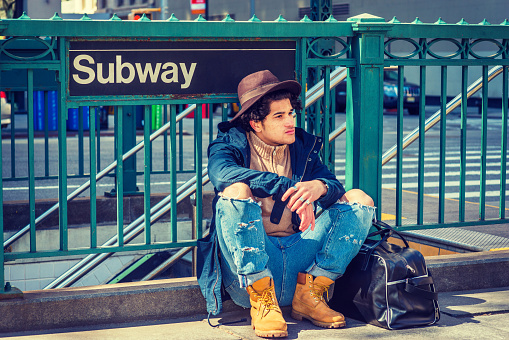 Young American Man with freckle face, curly hair, traveling in New York,  wearing blue jacket, jeans, yellow boot shoes, Fedora hat, black leather bag on ground, sitting on street by Subway sign.