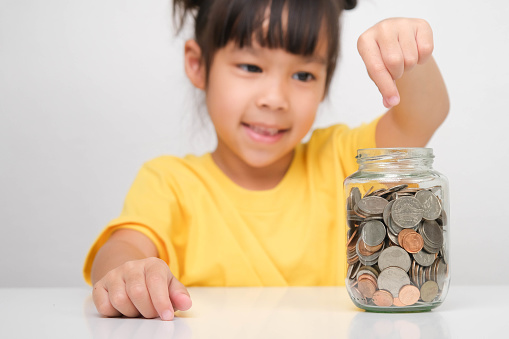 Happy smiling cute Asian girl holding a coin and putting it into a clear jar sitting at a table on a white background. Children learning about saving for future concept