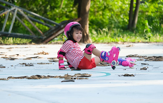Cute Asian little girl in protective pads and safety helmet practicing roller skating in the park. Exciting outdoor activities for kids. A preschooler wearing roller skates falls down.