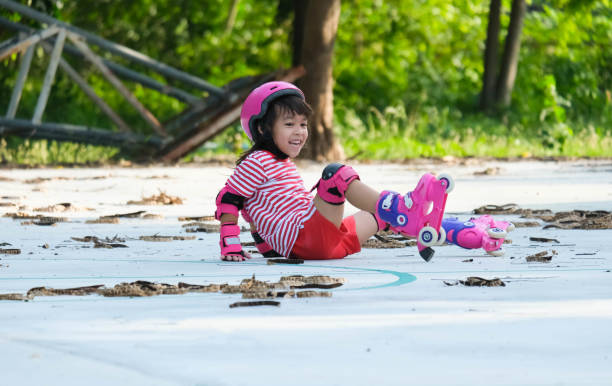 Cute Asian little girl in protective pads and safety helmet practicing roller skating in the park. Exciting outdoor activities for kids. A preschooler wearing roller skates falls down. Cute Asian little girl in protective pads and safety helmet practicing roller skating in the park. Exciting outdoor activities for kids. A preschooler wearing roller skates falls down. elbow pad stock pictures, royalty-free photos & images