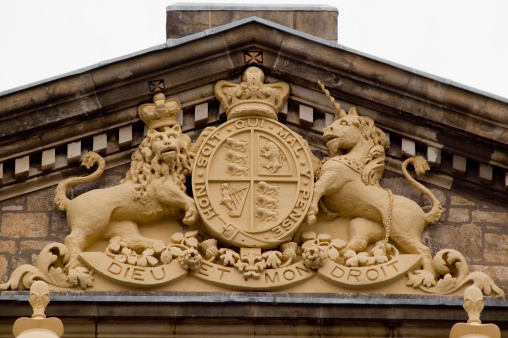 Yaren, Nauru: Coat of arms of the Republic of Nauru, tympanum of the Parliament building. The shield is divided and separated in the middle. In the upper section the alchemical symbol of phosphorus is shown over a woven background. The lower silver section depicts a black frigatebird, which sits on a perch over blue ocean waves. The lower right section is blue and contains a branch of calophyllum flowers. The shield is surrounded by images of tribal chief gear - ropes from palm leaves, feathers of the frigatebird and shark teeth. The twelve-pointed star above the shield is taken from the flag. The ribbon above it bears the name of the island in Micronesian Nauruan: \