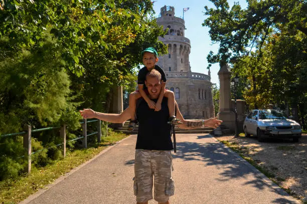 Photo of happy smiling son sitting on the shoulders of laughing father near the historic tower Elizabeth Lookout on Janos Hill. Budapest, Hungary