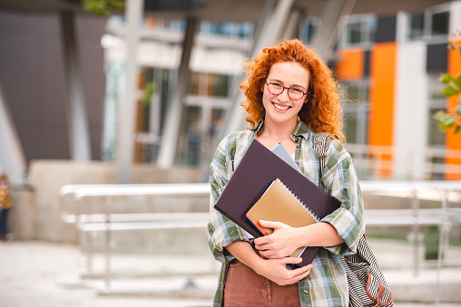 Beautiful Smiling Red Hair Student Girl Hugging Books Posing With Backpack Near College Building Outdoor. Modern Education, College Tuition And Grants, Studentship And Study Abroad Concept
