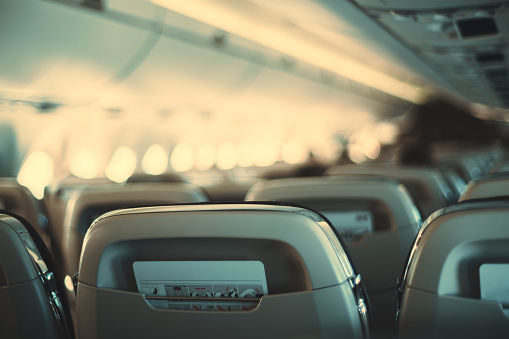 The long cabin of a passenger plane with a a shallow depth of field and selective focus on the closest empty row of seats with safety instructions in the pockets behind them, bokeh in aircraft windows