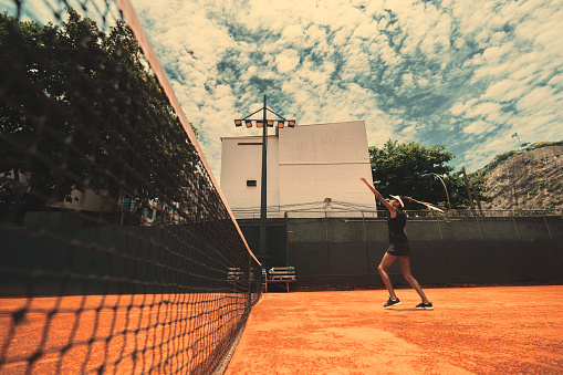 A wide-angle view from the corner of the tennis net of a slender black female in a sports outfit, on an orange clay ground of a private tennis club, swinging her racket to hit an incoming ball