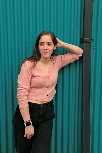 A Mexican woman leaning on a black downpipe against a teal wall. She is wearing a pink long sleeved top, black pants with earrings, ring and watch.