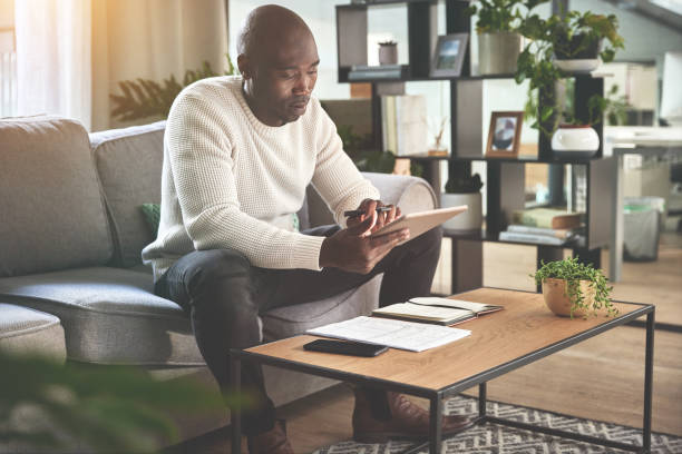 A man using a digital tablet and working with paperwork at home. A young man paying bills, planning his finance online with a touchscreen on the sofa