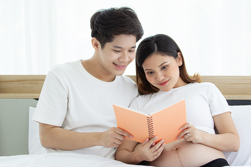 Portrait of lovely husband and wife reading a book together on the bed in bedroom, husband embracing his pregnant wife on a bed during reading a book. Couple relaxing together. Family leisure activity