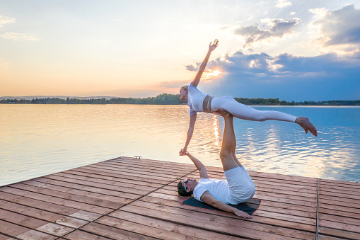 Beautiful young couple doing acro yoga on the pier against sunset. Man lying on the pier and balancing woman in his feet.