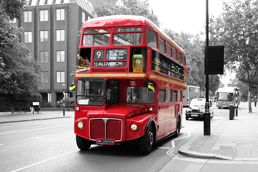 A traditional red double-decker bus isolated on a black and white background. The bus is on a moderately busy street in London and is directly followed by a black cab is a tourist coach. The picture is taken under a three-quarter angle from back right to front left.