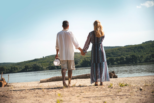 Rear view on Mature Couple walking on sunny day outdoors.  Man and Woman holding hands in walk on sand beach by the river. Couple in love enjoying nature. Copy space