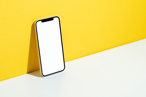 Blank white screen smart phone mockup, template with clipping path on yellow and white background. Phone leaning against to yellow wall.
