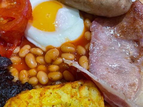 Stock photo showing an elevated view of a white plate containing a Full English breakfast with bacon rasher, grilled sausage, baked beans, half a grilled tomato, hash brown and fried egg.