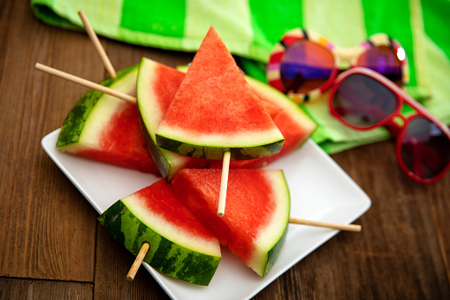 This is a high angle photograph of a plate of triangle shaped sliced cut watermelon with popsicle sticks making it easier to eat on a white modern plate. It is surrounded by sun glasses a beach towel and sitting on a wood Beach deck