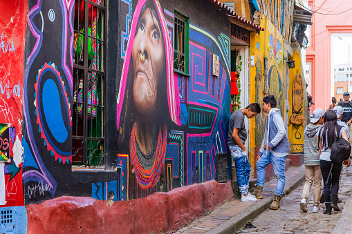 Bogota, Colombia - July 20, 2016: Both Tourists and local Colombian people walk up the narrow Calle del Embudo one of the most colorful streets in the historic La Candelaria district of Bogotá, the Andean capital city of the South American country of Colombia. The street leads to the Chorro de Quevedo, the plaza where it is believed the Spanish Conquistador, Gonzalo Jiménez de Quesada founded the city in 1538. Many street facing walls in this area are painted with either street art or the legends of the pre-Colombian era, in the vibrant colours of Colombia. The altitude at street level is 8,660 feet above mean sea level. Photo shot on an overcast morning; horizontal format.