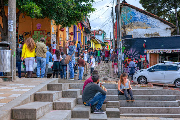 Bogotá, Colombia - Local Colombians And Tourists On The Cobblestoned Calle del Embudo, In The Historic La Candelaria District of The South American Andes Capital City Bogotá, Colombia - May 28, 2017: A few tourists and some local Colombians seen on the broader end of the, colourful, cobblestoned Calle del Embudo which is at the opposite end of the Plaza del Chorro de Quevedo. The Street gets its name from its funnel shape: Embudo translates to Funnel in English. It is shaped like a funnel. These streets were set up over 450 years ago when people usually travelled on horseback. The Street is well known for its Street Art, many of which feature the legends of the Pre-Colombian era. Photo shot in the late afternoon sunlight; horizontal format. calle del embudo stock pictures, royalty-free photos & images
