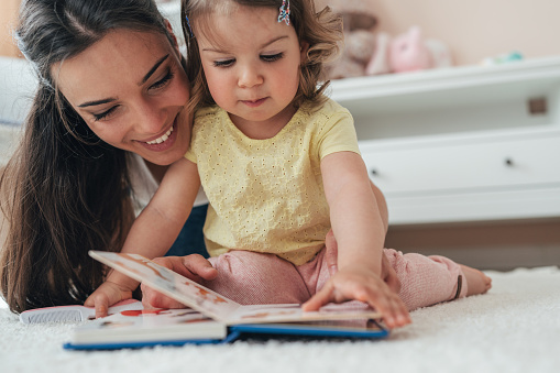 Young beautiful mother reading book with toddler daughter on the floor at home